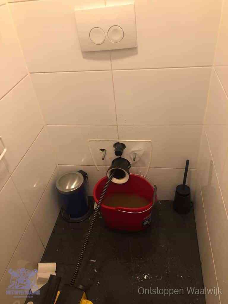 Een ontstopte wand-wc met verwijderde toiletpot waar een ontstoppingsveer in de wanduitgang verdwijnt, het achterhalen waar de verstopping zit in Waalwijk