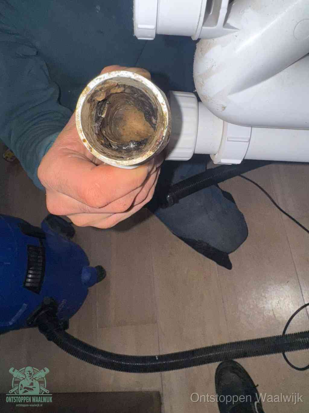 Een hand houdt een losgekoppeld PVC-afvoerbuisstuk vast dat van binnen volledig is dichtgeslibd met vet, vuil en aanslag, de meest voorkomende oorzaak van een verstopte afvoer in Waalwijk