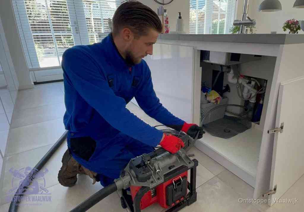 Een monteur voert een professionele elektrische ontstoppingsmachine in de afvoer onder de spoelbak tijdens het gootsteen ontstoppen in Waalwijk